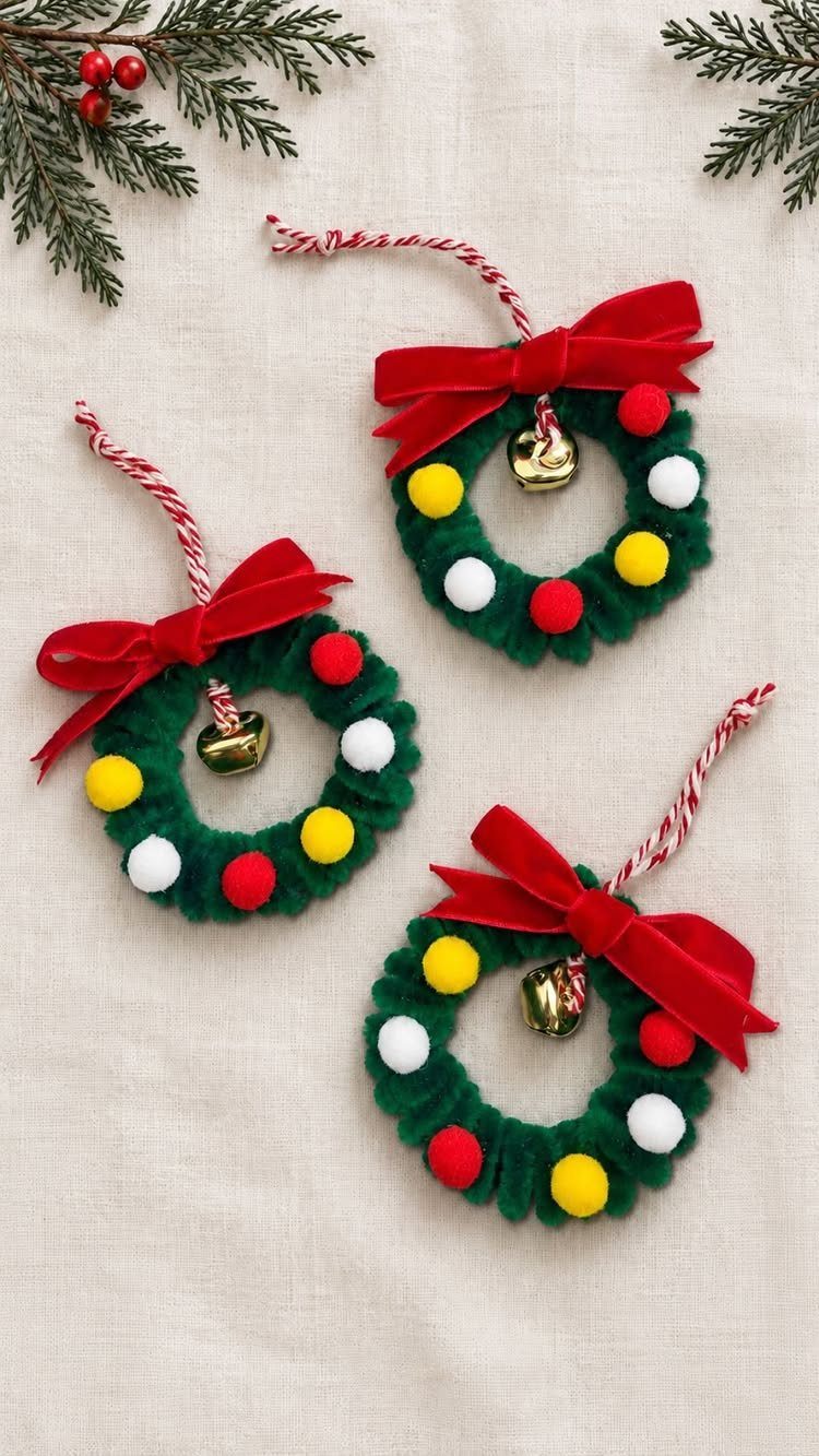 Three green wreath ornaments with red bows, gold bells, and striped twine hanging loops on fabric.