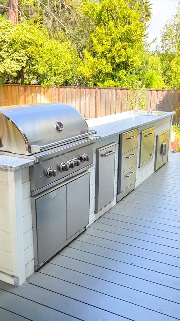 A stainless steel built-in outdoor grill station with stone countertops on a grey wooden deck.