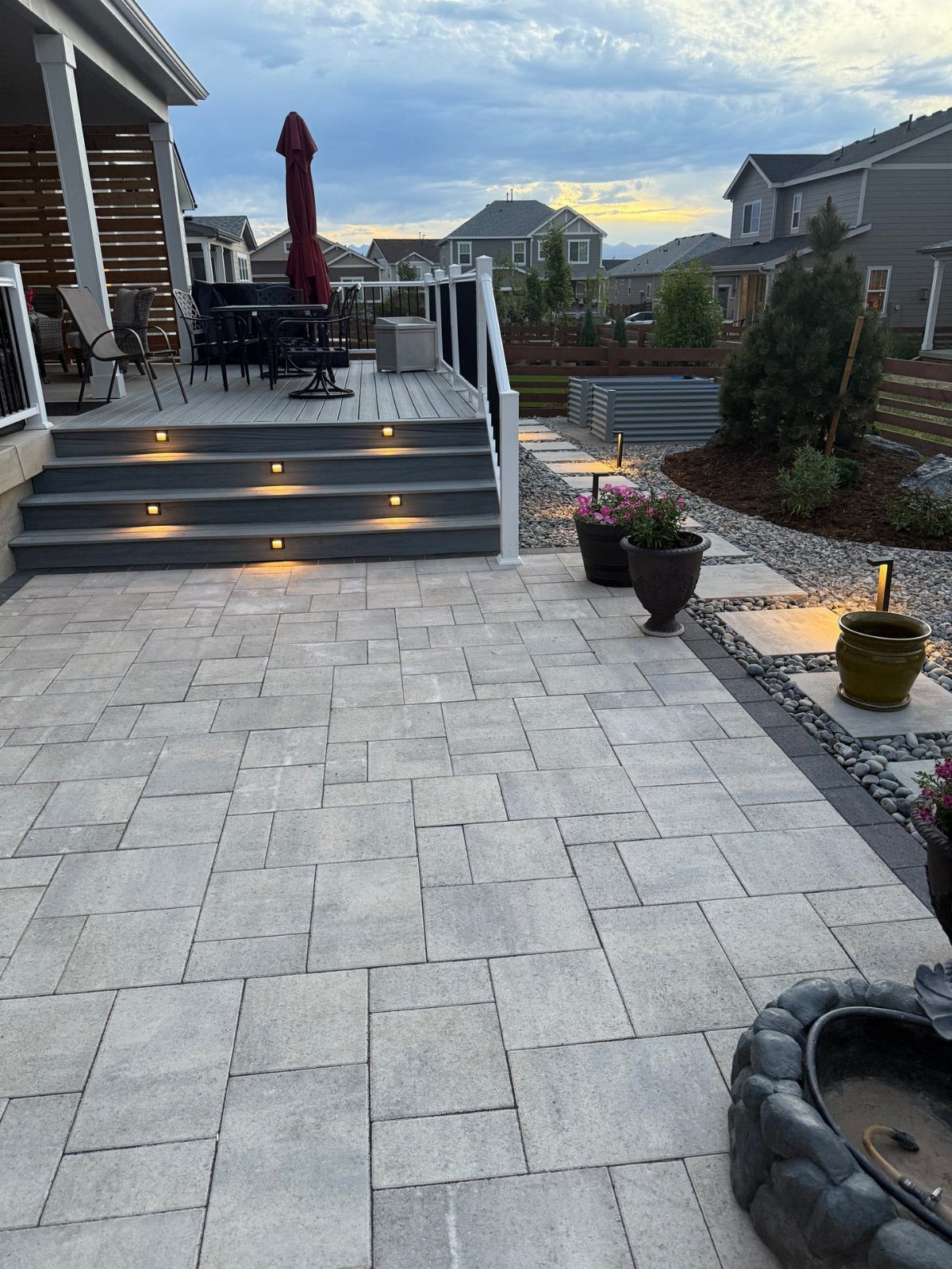 Grey wooden deck with lit steps connecting to a large stone paver patio in a backyard.