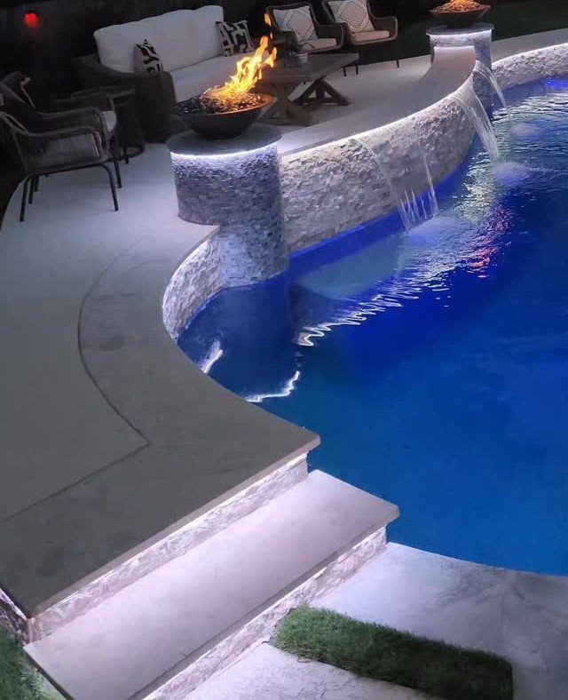 A glowing blue swimming pool with a stone waterfall feature and a fire pit nearby.