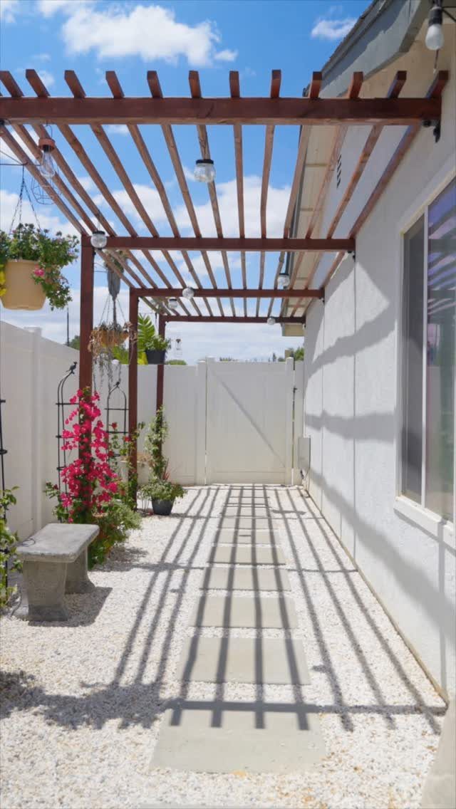 Wooden pergola casting striped shadows over a gravel walkway with stone pavers and pink flowers.