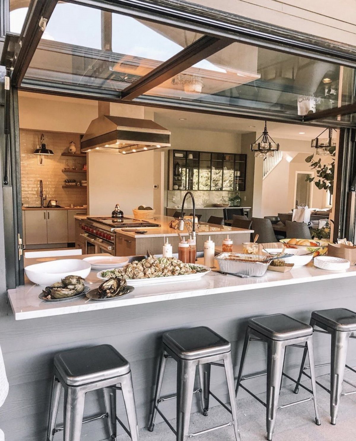 Barndominium Indoor Outdoor Kitchen: 8 Ideas That Erase the Wall 3 Black-framed folding glass window opening a modern kitchen to an outdoor bar with metal stools.