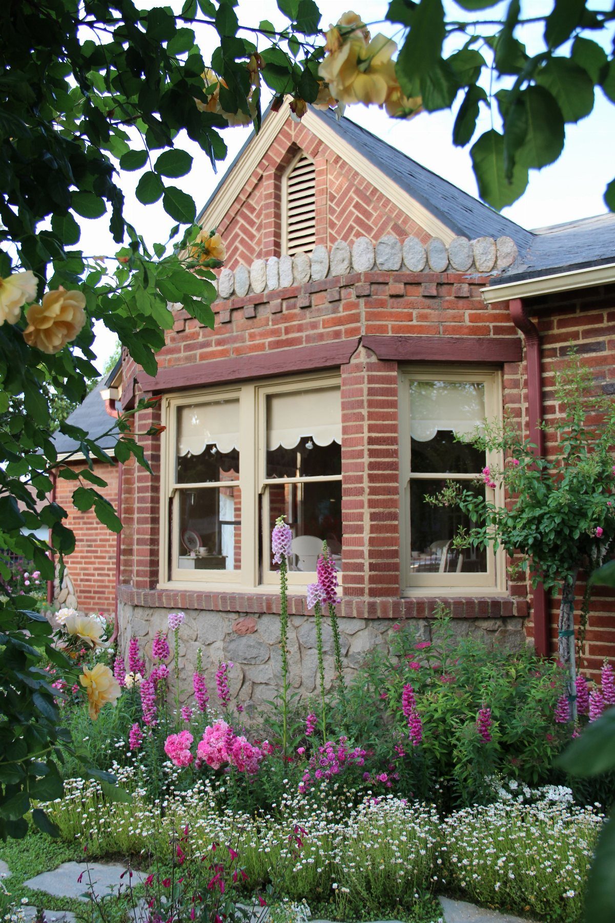 Pink foxgloves and white daisies bloom in a lush garden in front of a brick bay window.