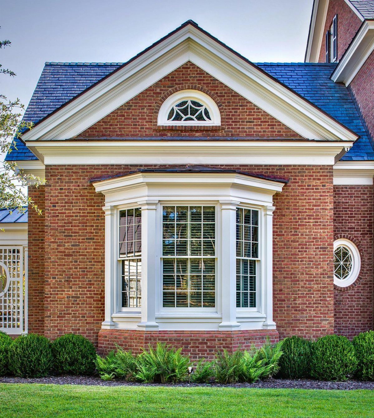 A red brick house featuring a white bay window with decorative moldings and green shrubs below.