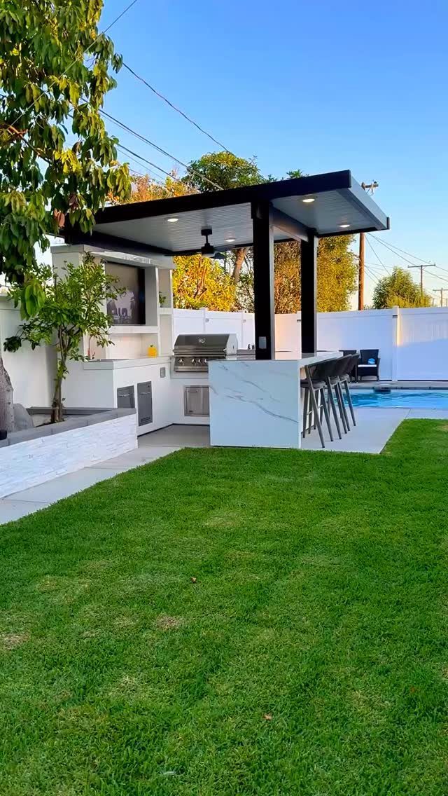 Modern built-in outdoor kitchen with marble counters, grill, and black pergola next to a lawn.