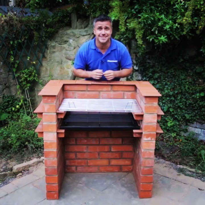 A man standing behind a large, handmade red brick outdoor barbecue structure on a patio.