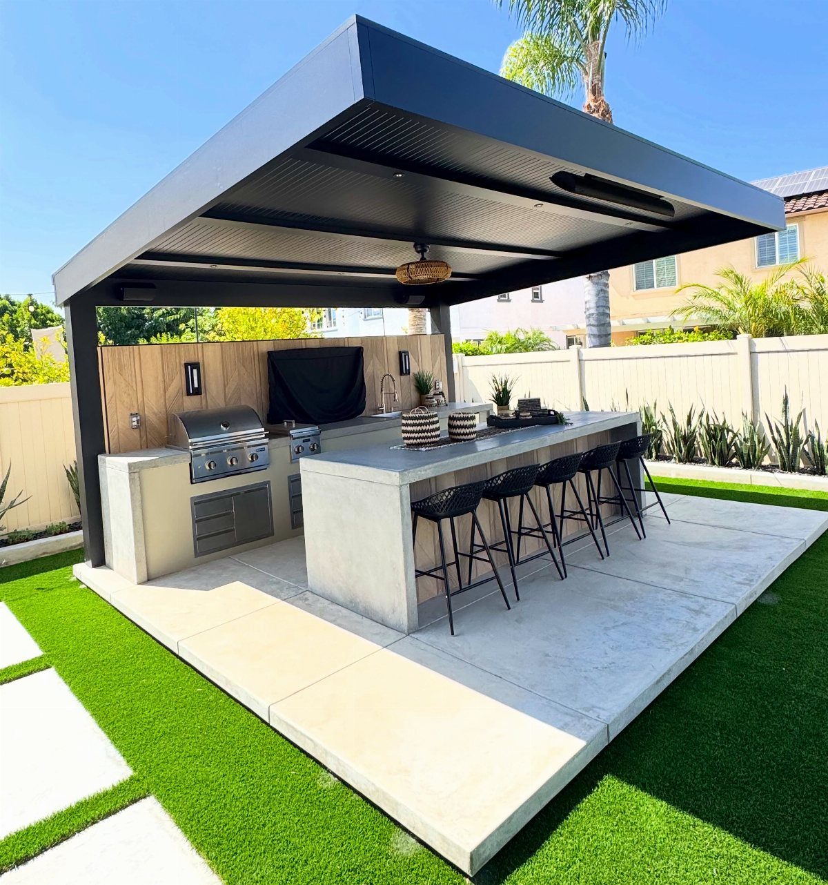 Modern outdoor kitchen with a built-in grill and concrete island under a black metal pergola.