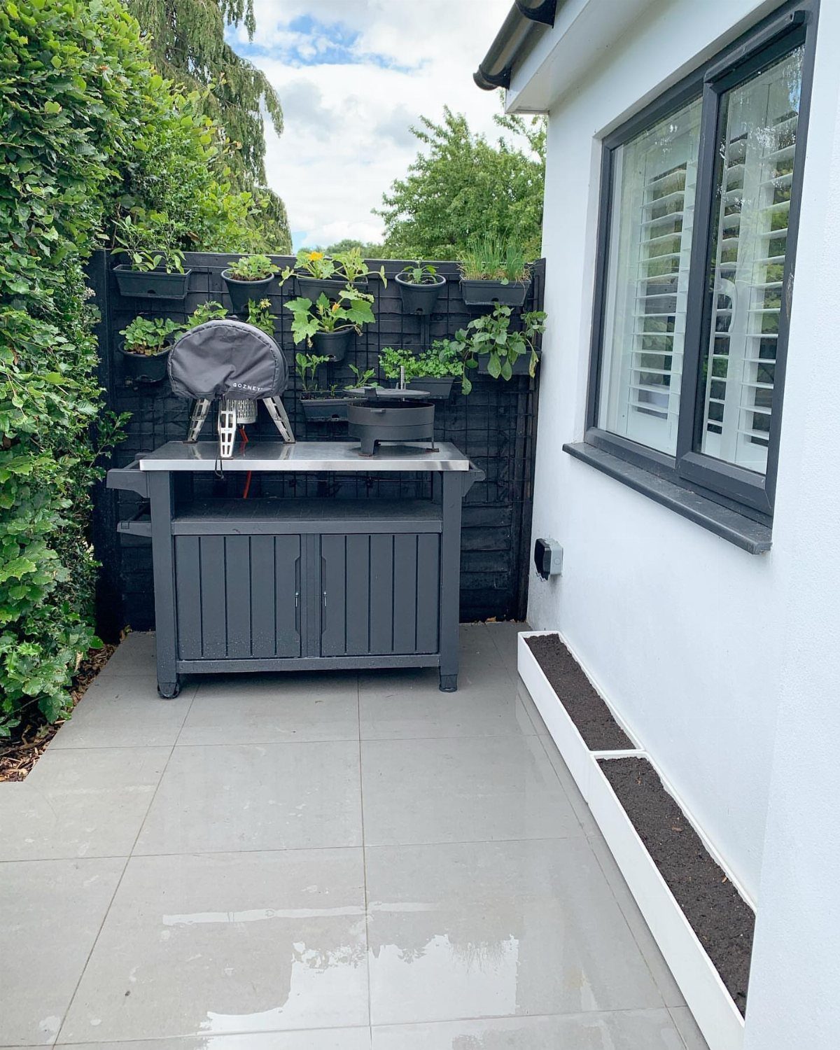 Black BBQ cart and vertical planters on a grey tiled patio next to a white house.