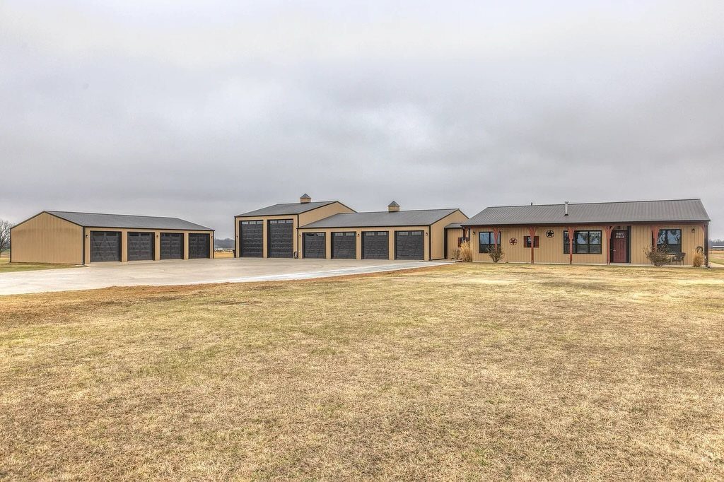 Three large tan metal buildings with dark roofs and multiple garage doors on a grassy field.