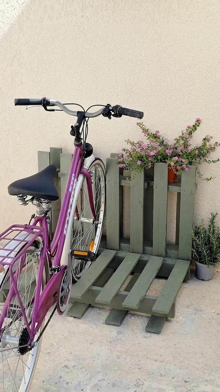 A purple bicycle parked next to a green wooden pallet bike stand against a wall.