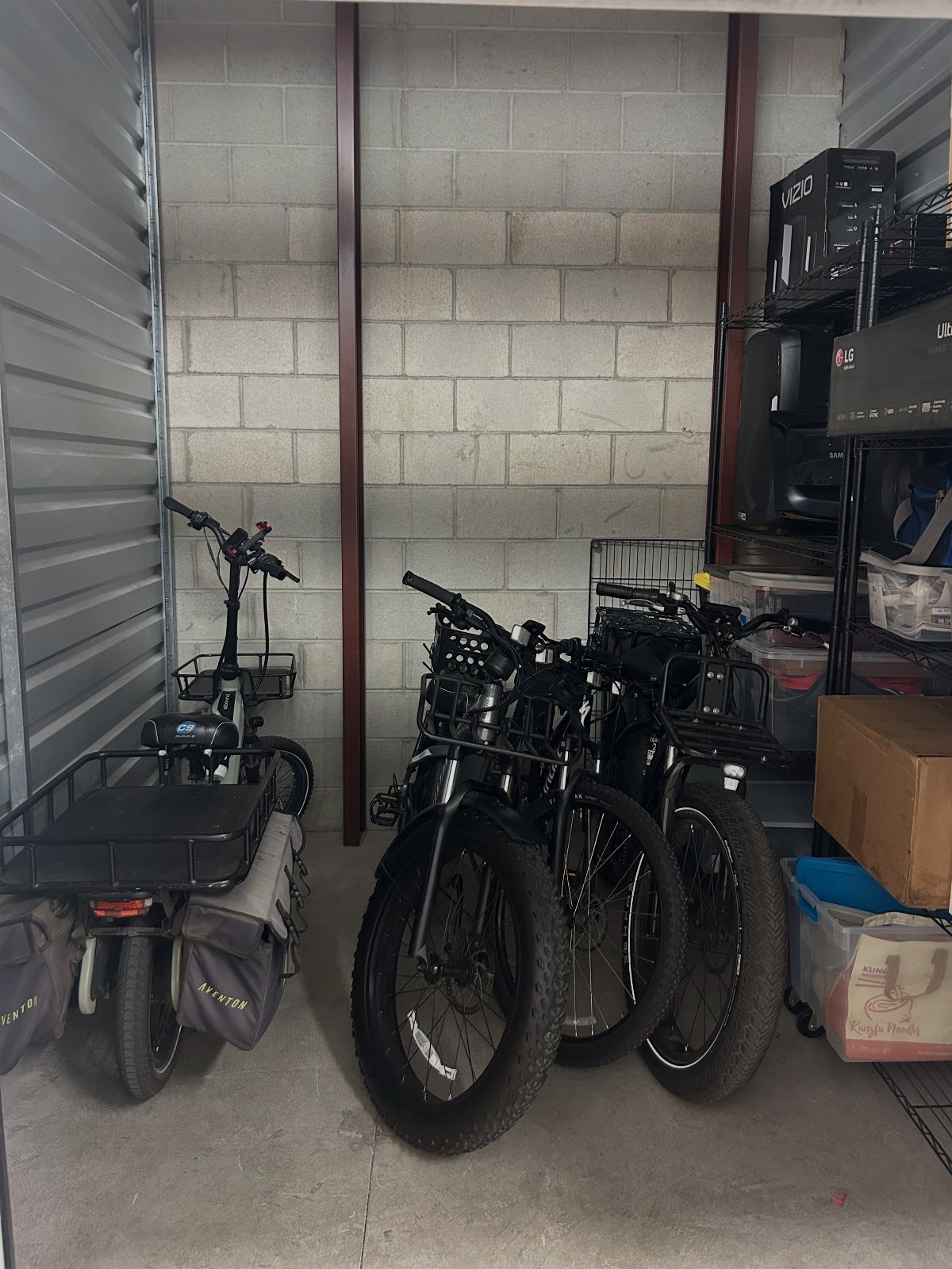 Two black electric bikes parked inside a cinder block storage unit next to metal shelving.