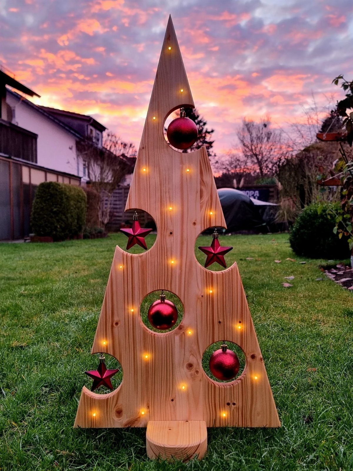 25 Christmas Art and Craft Ideas for Cards, Ornaments, and Decor 10 A wooden shim Christmas tree with red ornaments and glowing lights on a lawn.