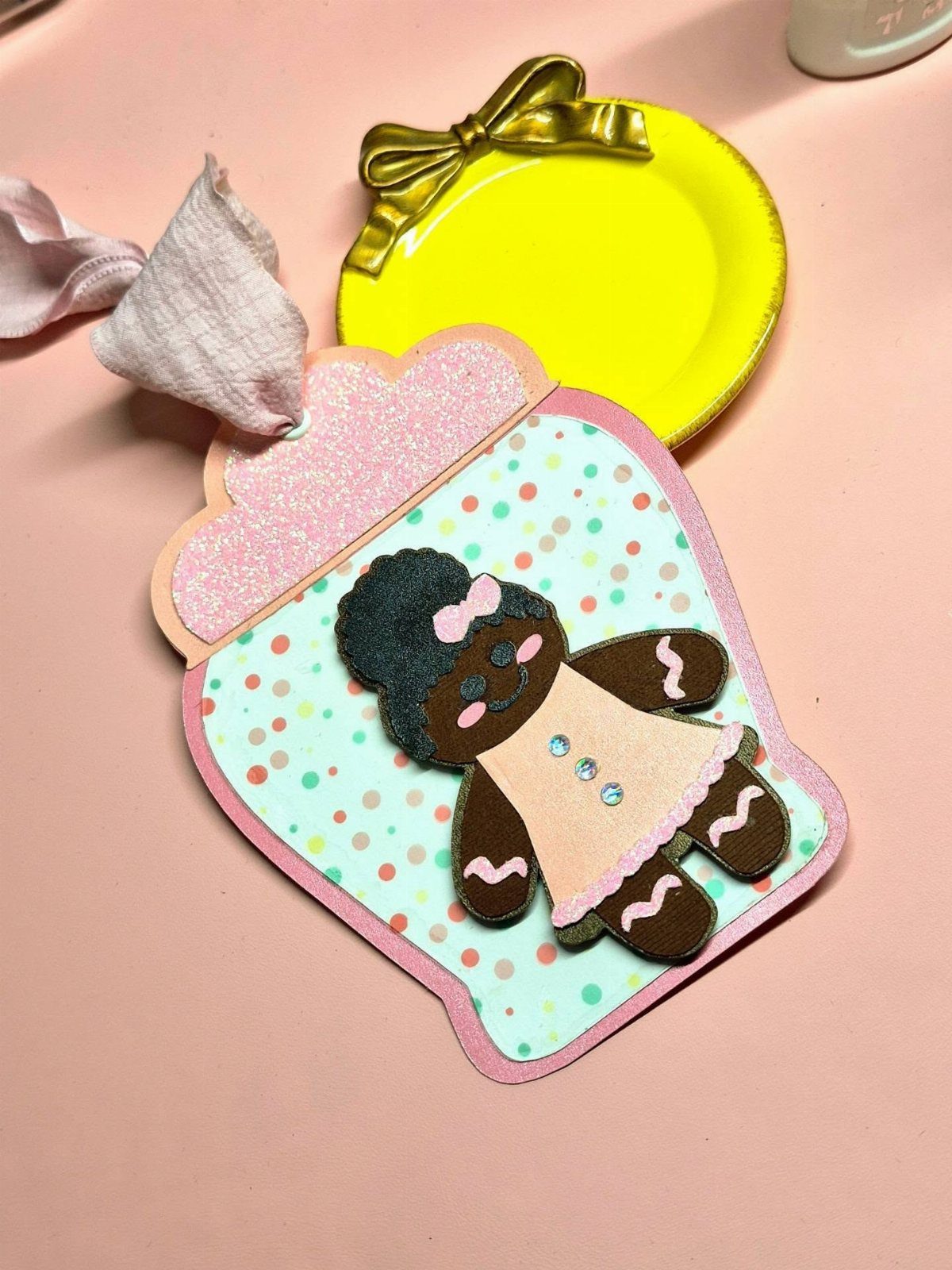 25 Christmas Art and Craft Ideas for Cards, Ornaments, and Decor 17 A handmade gingerbread girl paper gift tag with pink glittery edges on a pink surface.