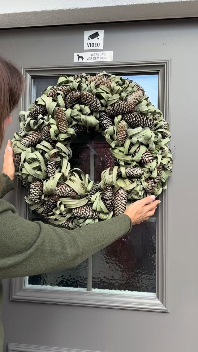 25 Christmas Art and Craft Ideas for Cards, Ornaments, and Decor 15 A person holding a large sage green and pinecone wreath against a grey door.