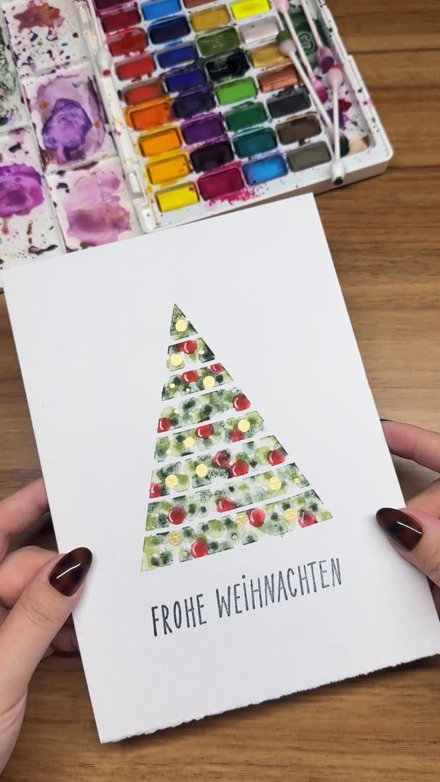 25 Christmas Art and Craft Ideas for Cards, Ornaments, and Decor 4 Hands holding a handmade watercolor Christmas card with a decorated tree design on a wooden table.
