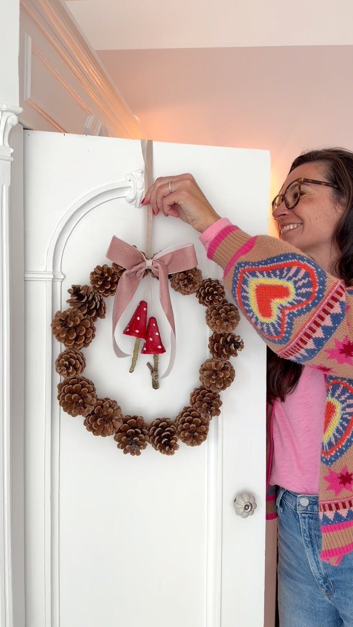 Christmas Decorations with Pinecones: 17 Ideas for Your Home 2 A woman hanging a pinecone wreath decorated with a pink bow on a white door.