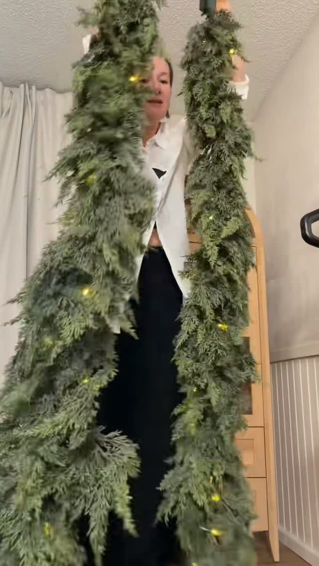 5 Christmas Stairs Decor Ideas: Garland, Bells, Paper Orbs & More 1 A person holding two long green evergreen garlands decorated with warm yellow fairy lights.