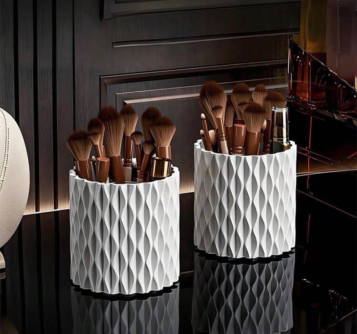 Two white textured containers filled with various makeup brushes on a dark reflective surface.