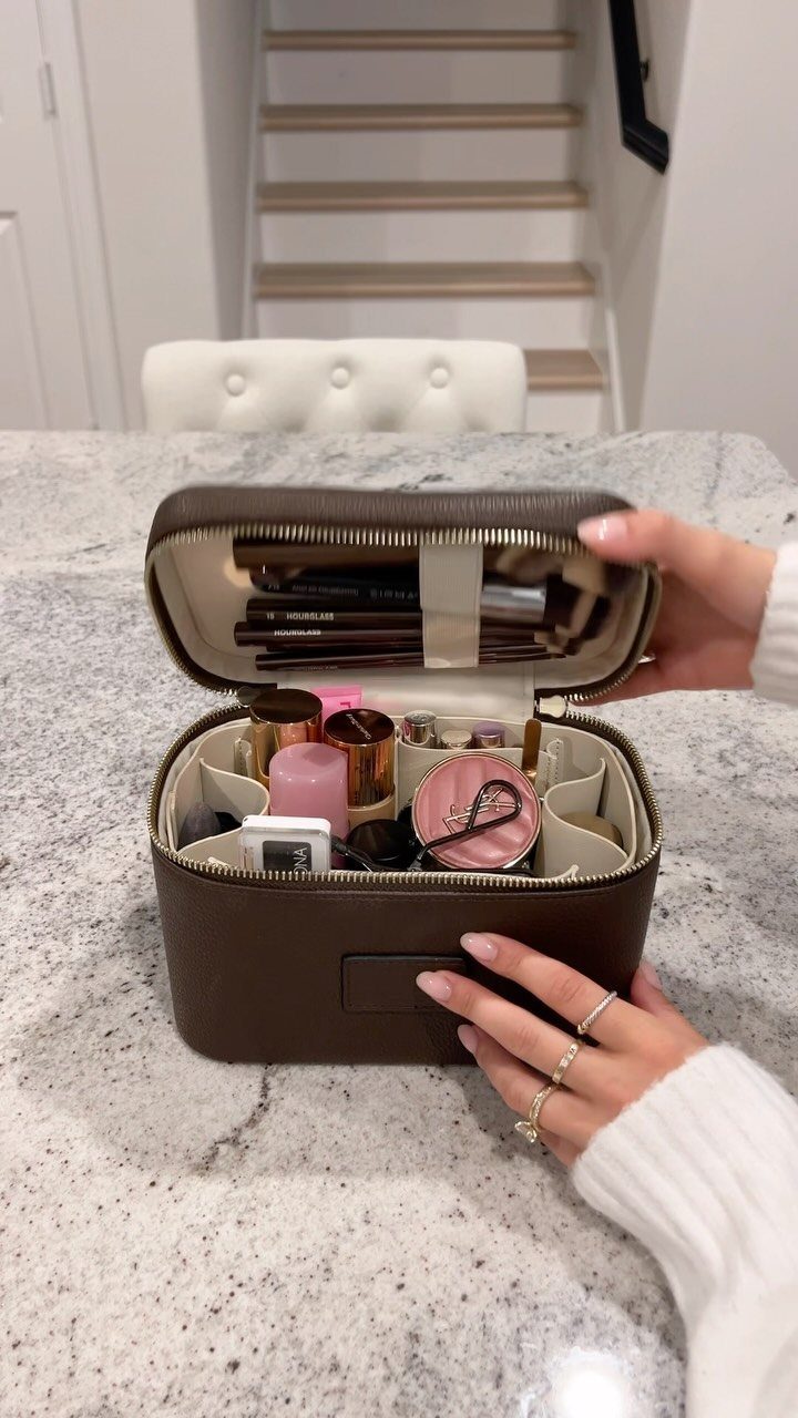 Hands holding open a brown leather makeup case filled with organized cosmetics on a countertop.