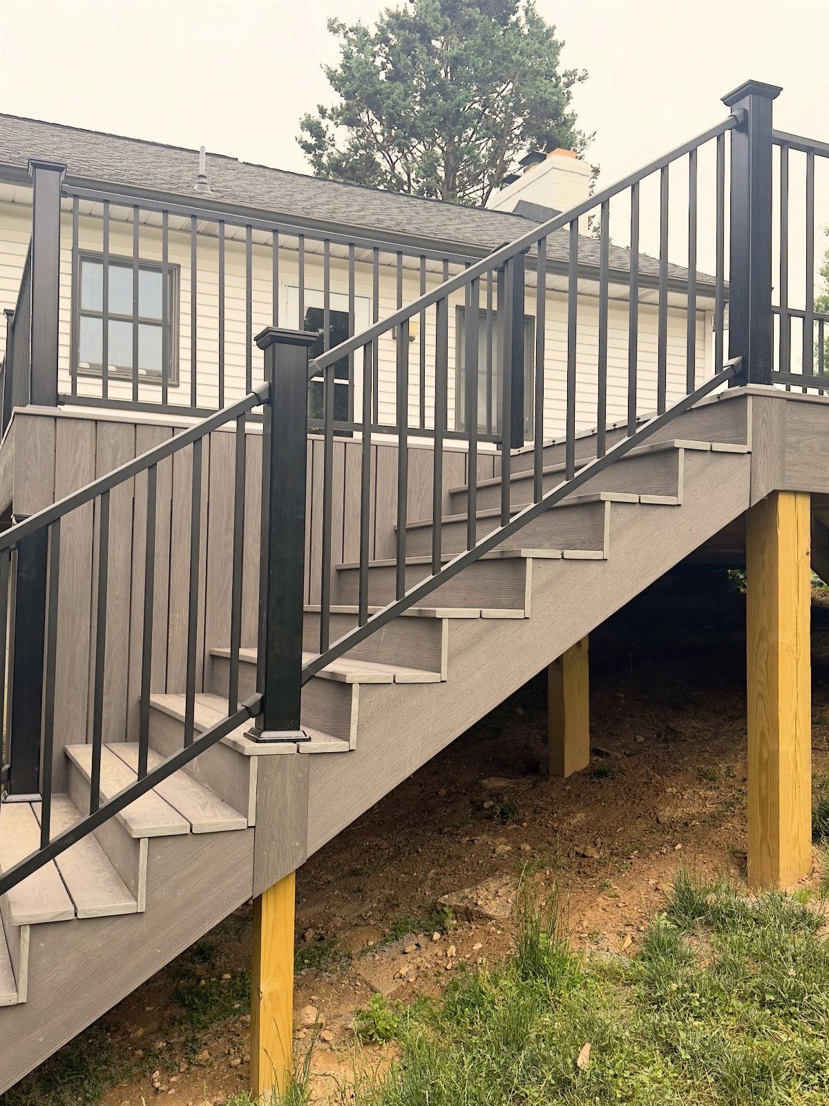 Deck on a Slope: 5 Ideas That Beat the Grade 3 Grey composite deck stairs with black metal railings descending a grassy slope on wooden posts.