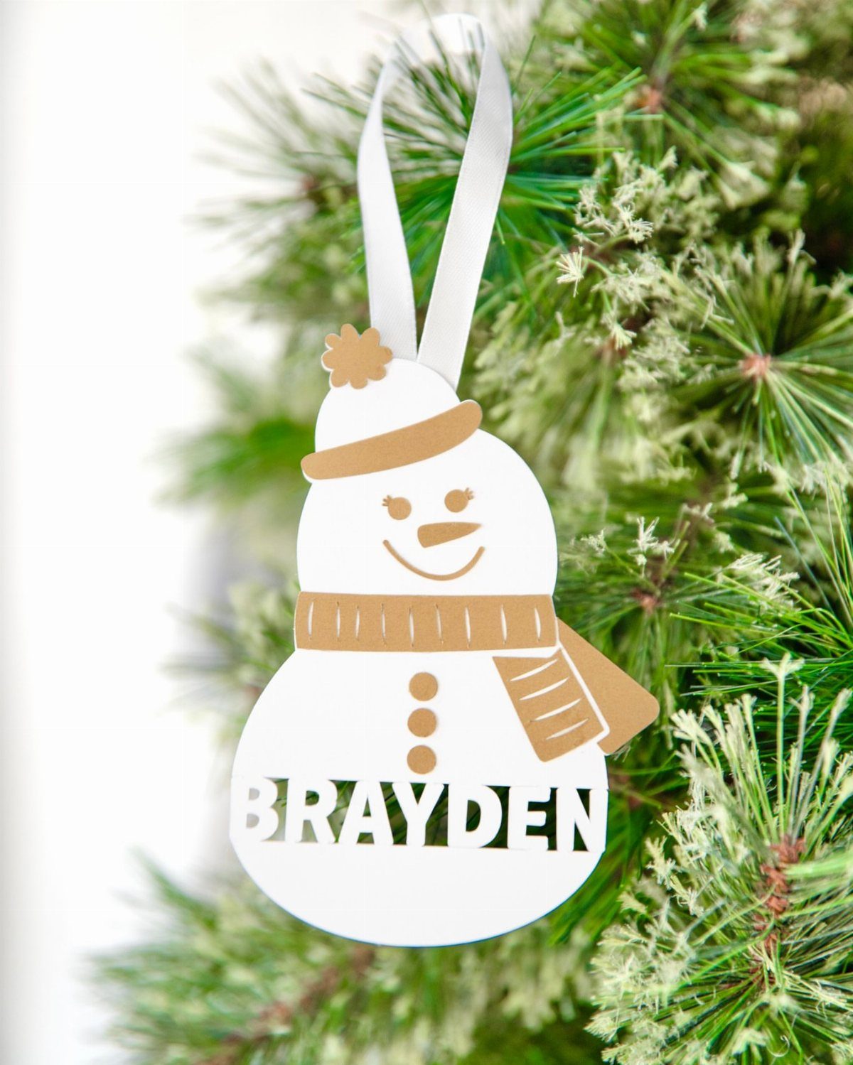 21 DIY Christmas Ornaments for Adults 8 A white snowman ornament with tan details and the name Brayden hanging on a pine branch.