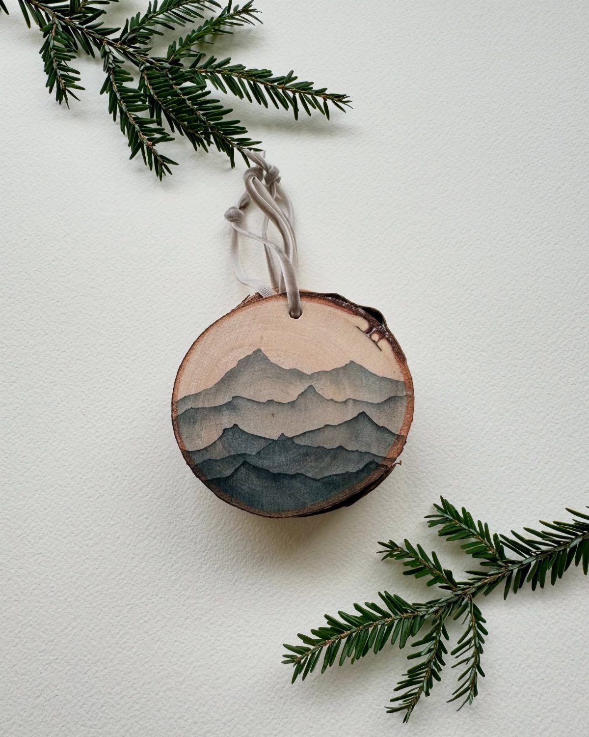 21 DIY Christmas Ornaments for Adults 6 Painted birch wood slice ornament featuring a blue mountain landscape hanging between evergreen branches.