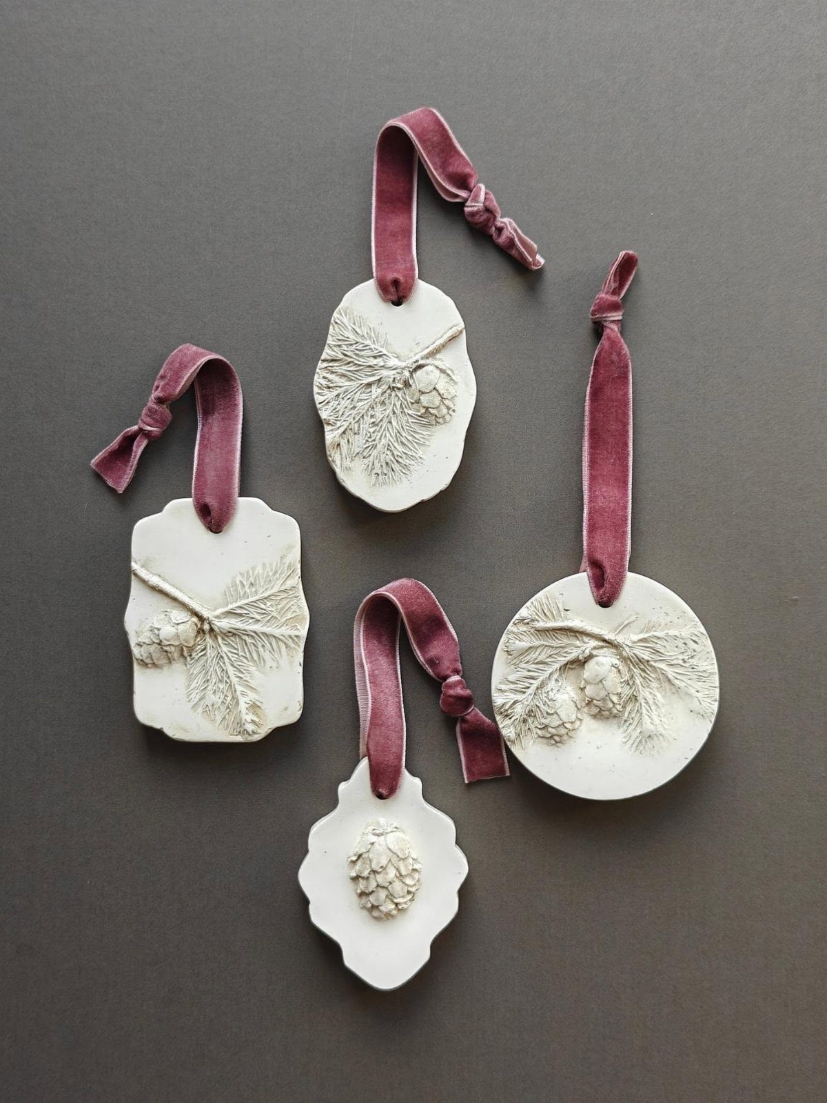 Easy Christmas Clay Ideas: 9 Projects for Handmade Ornaments 9 Four white clay ornaments with pine needle impressions and burgundy velvet ribbons on a dark background.