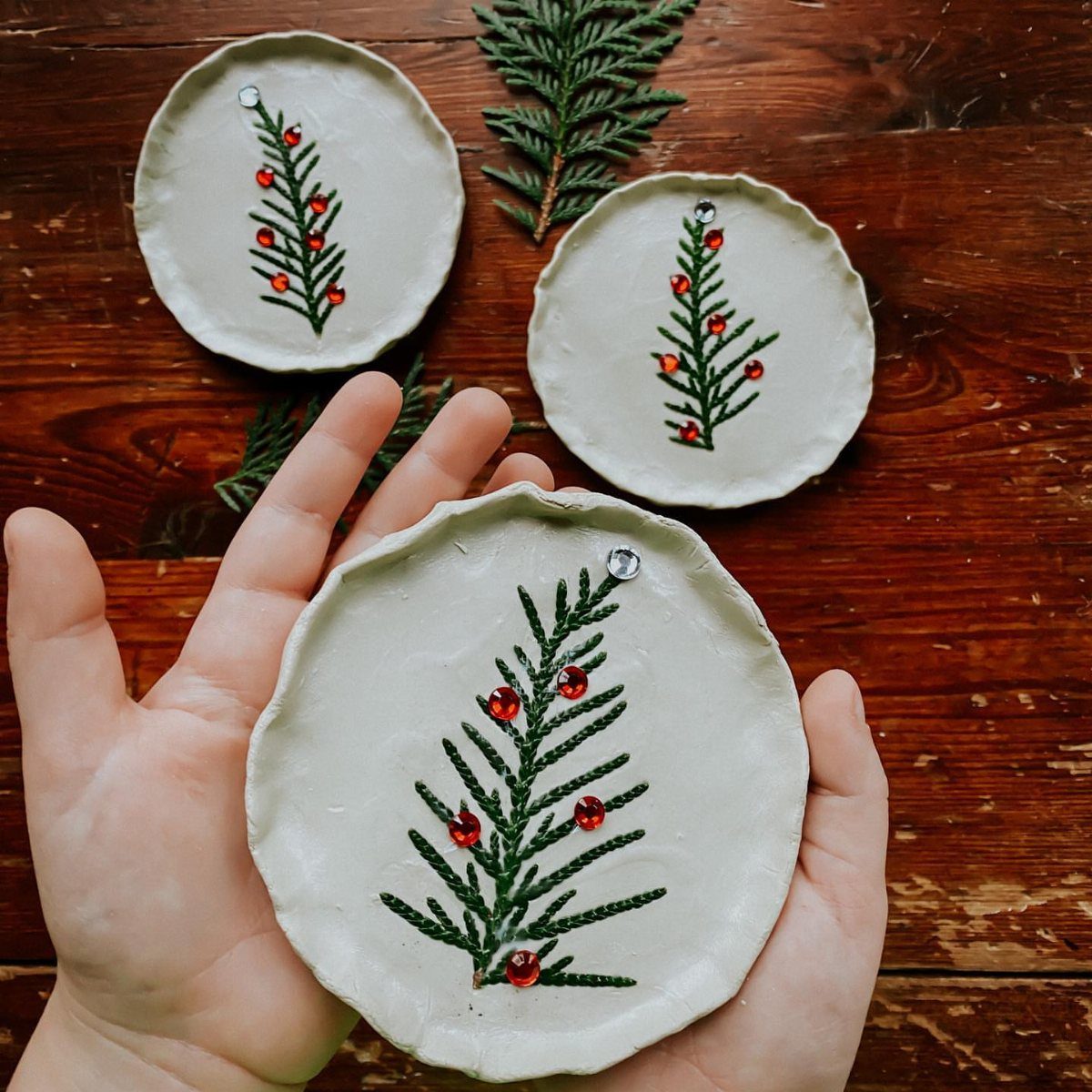 Easy Christmas Clay Ideas: 9 Projects for Handmade Ornaments 7 Hands holding a white clay ornament with a green pine needle imprint and red decorations.