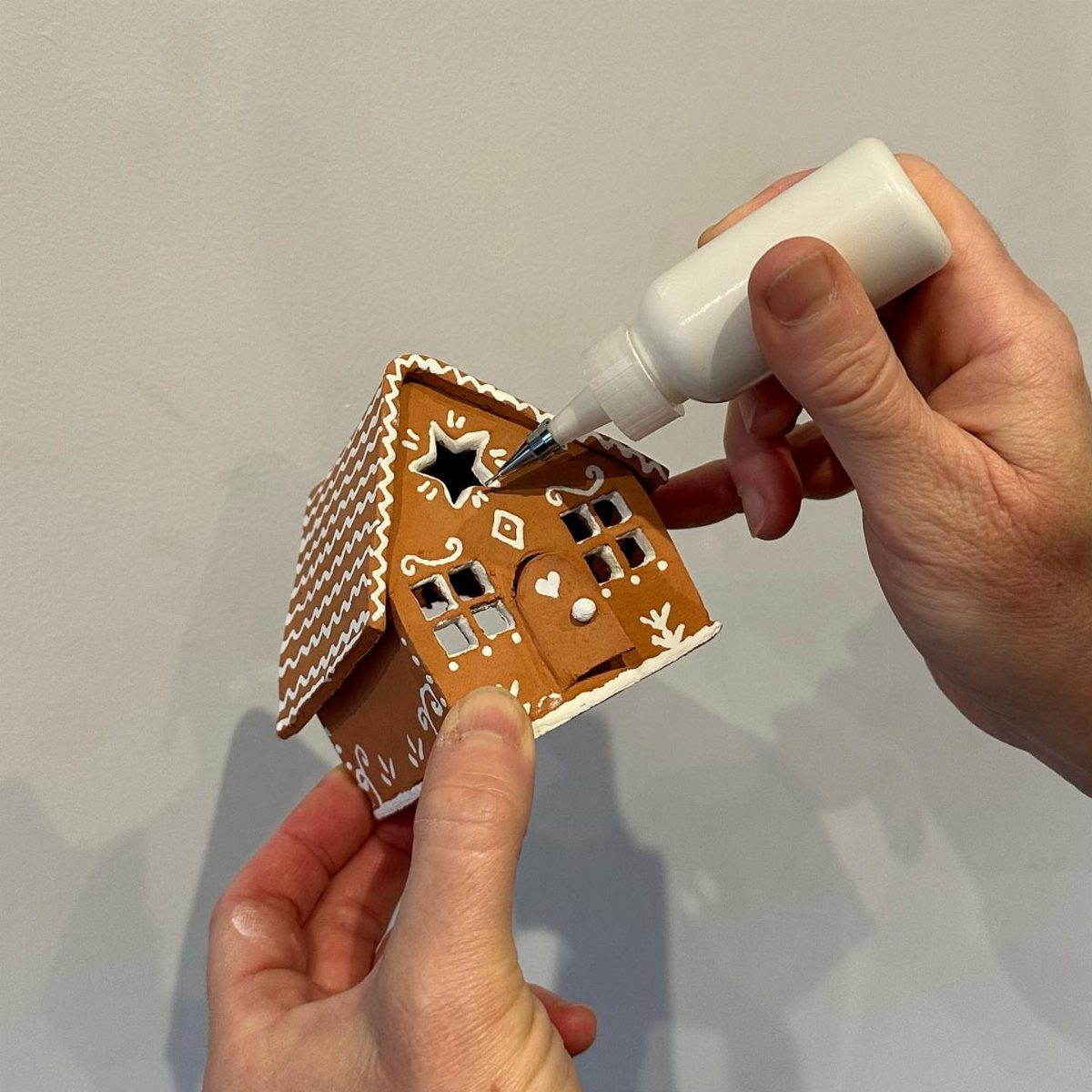 Easy Christmas Clay Ideas: 9 Projects for Handmade Ornaments 8 Hands using a white squeeze bottle to decorate a small brown clay gingerbread house.