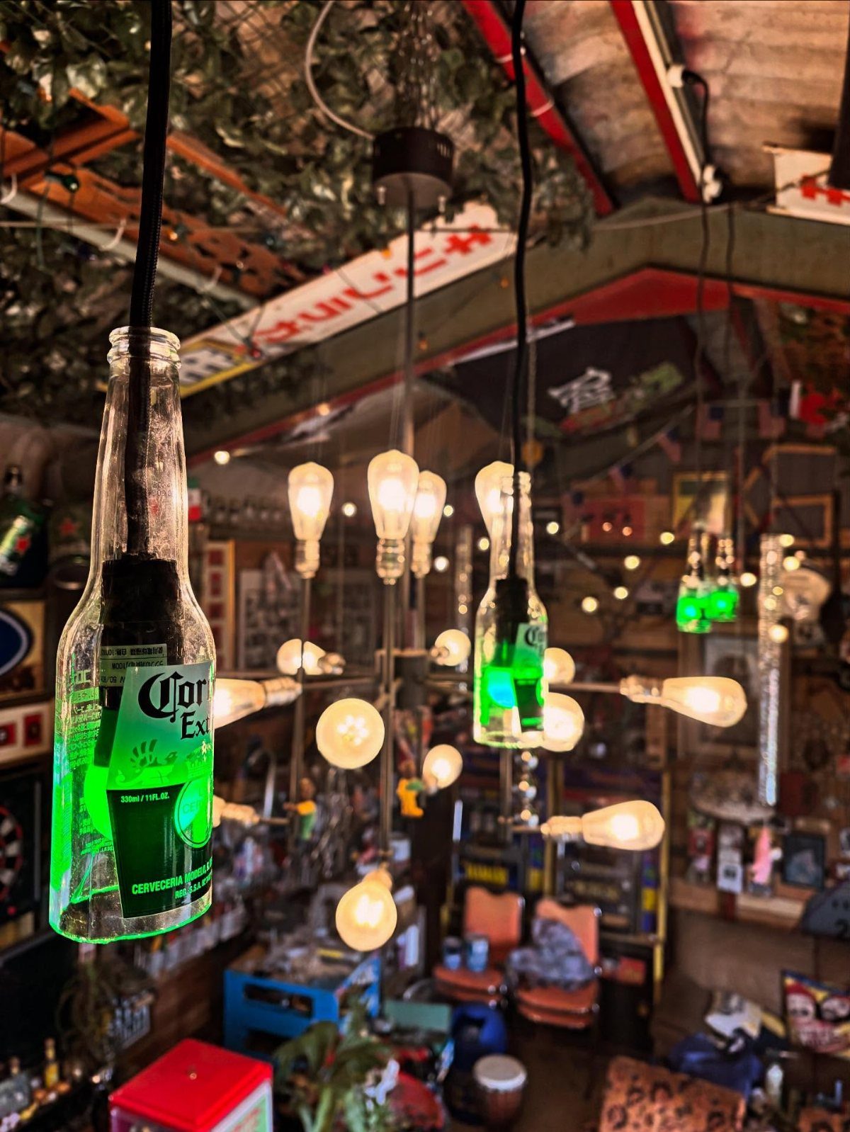 Repurposed beer bottle pendant lights glowing green amidst warm hanging Edison bulbs in a dim room.