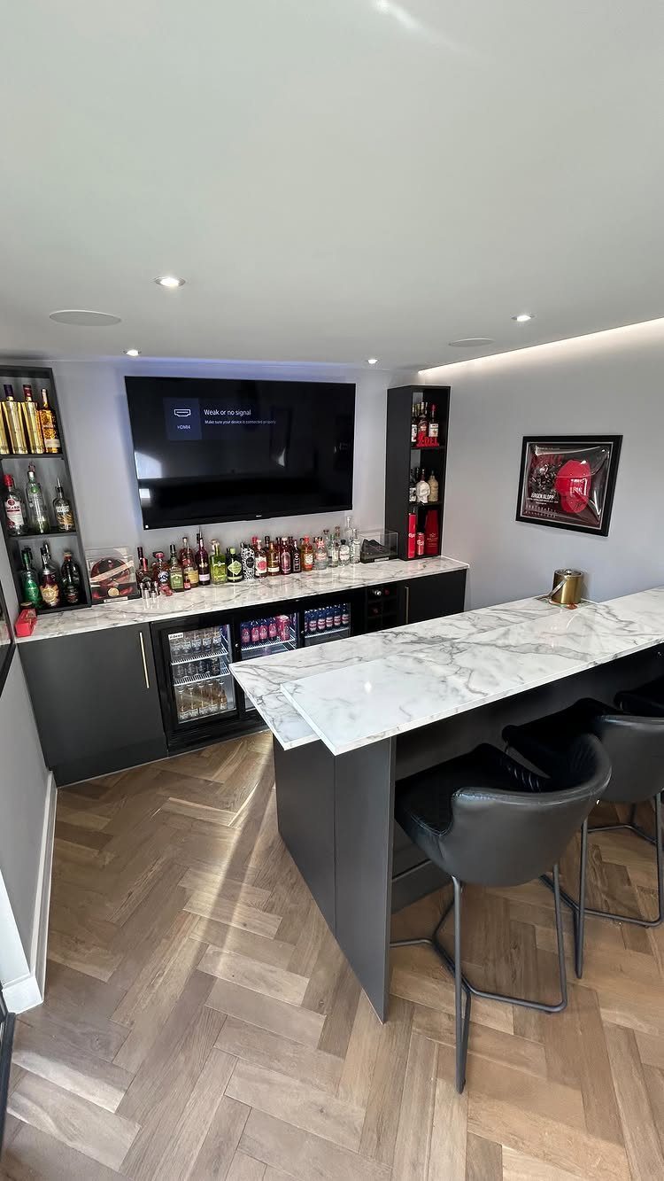 Modern home bar with marble countertops, liquor bottles, and light wood herringbone flooring.