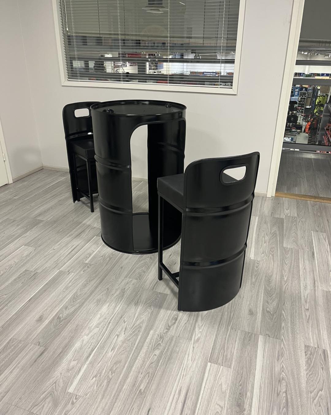 Black metal barrel-style bar table and two matching stools on a grey floor.