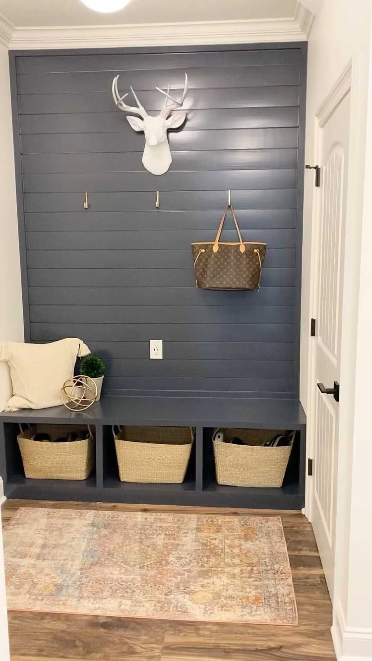 Dark blue shiplap wall with gold hooks, a deer head mount, and a bench with baskets.