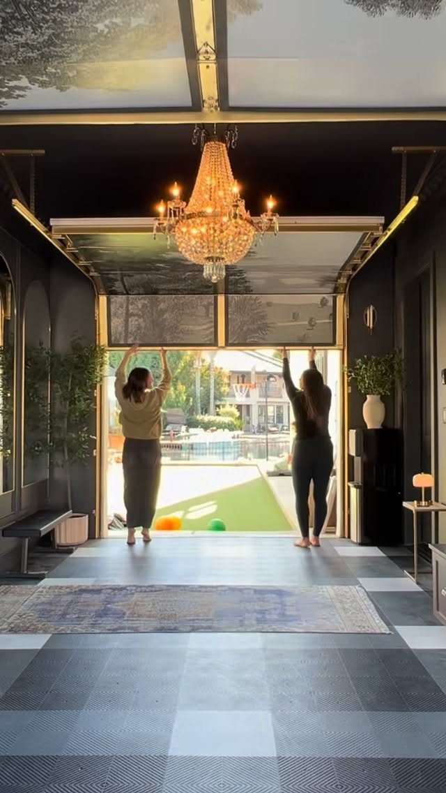 Two women opening large glass doors in a dark, modern entryway featuring a patterned area rug.