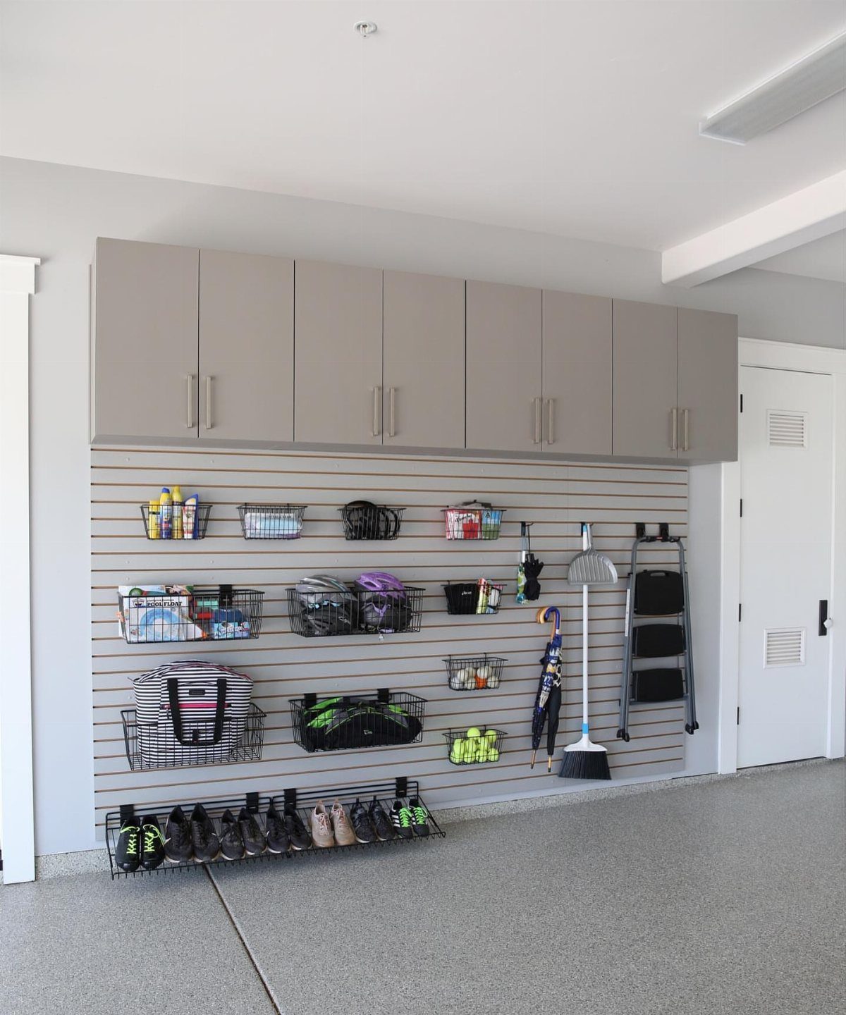 A grey slatwall storage system with wire baskets and tools mounted below grey cabinets in a garage.