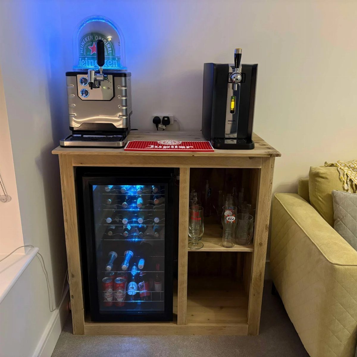 10 Garage Hangout Ideas for Comfortable Seating, Lighting, and More 7 Wooden bar unit with a mini fridge, beer dispenser, and beverage machine against a white wall.