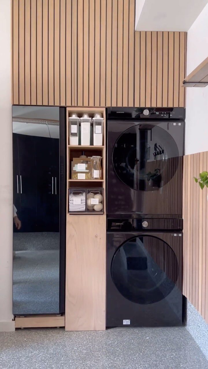 Stacked black washer and dryer next to a refrigerator and wooden storage shelves.