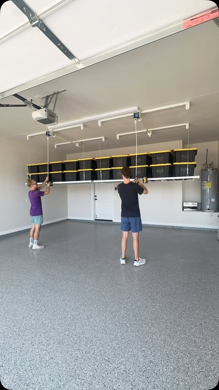 Two people installing a ceiling-mounted storage rack holding black bins in a garage.