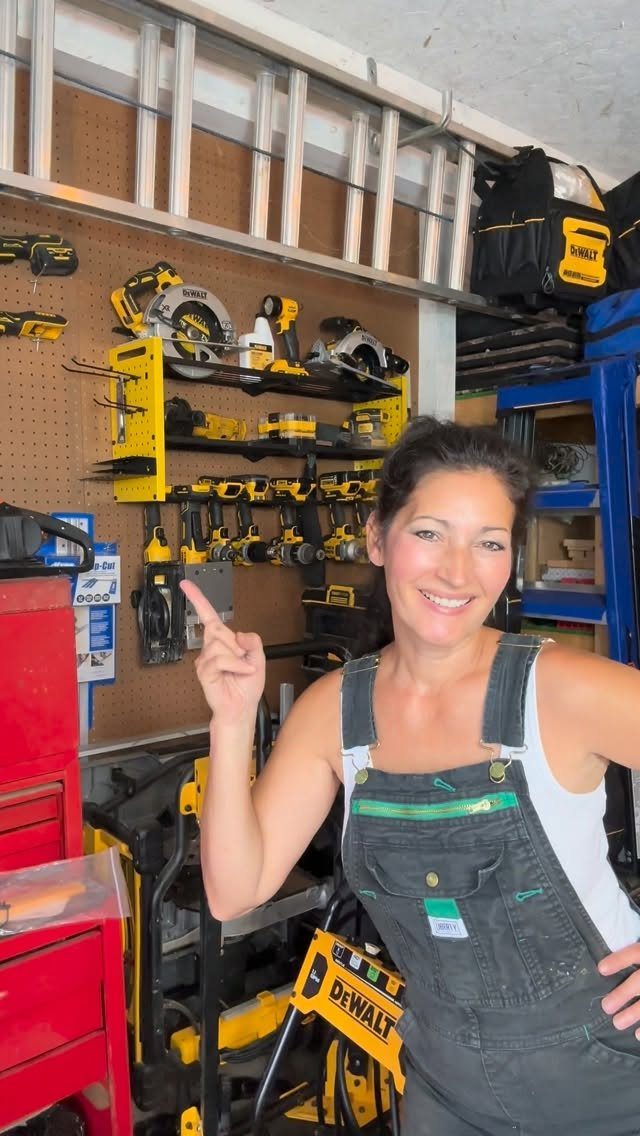 Smiling woman in overalls pointing at organized power tools on a garage pegboard wall.