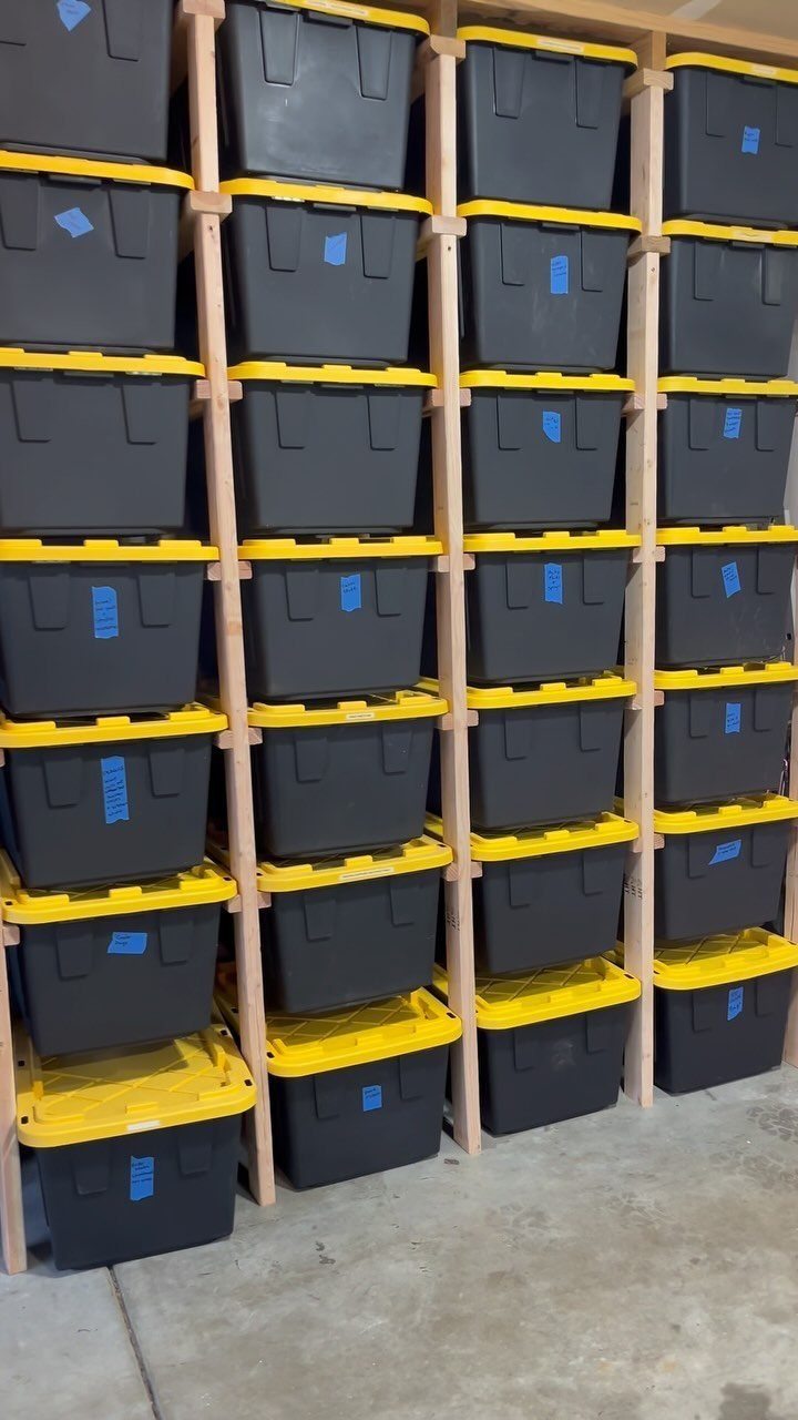 Black storage bins with yellow lids stacked on wooden shelves in a garage.