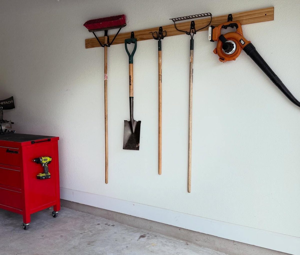 Garden tools like a shovel and rake hanging from hooks on a wooden strip against a white wall.