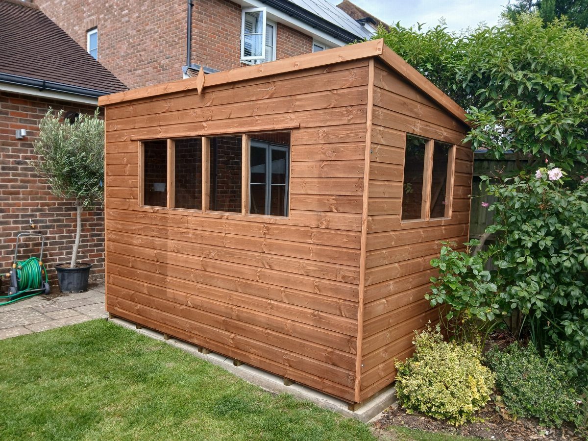 Garden Shed Design: 7 Ways to Use the Space You Have 4 A wooden pent shed with a sloped roof situated in a green garden next to a brick house.