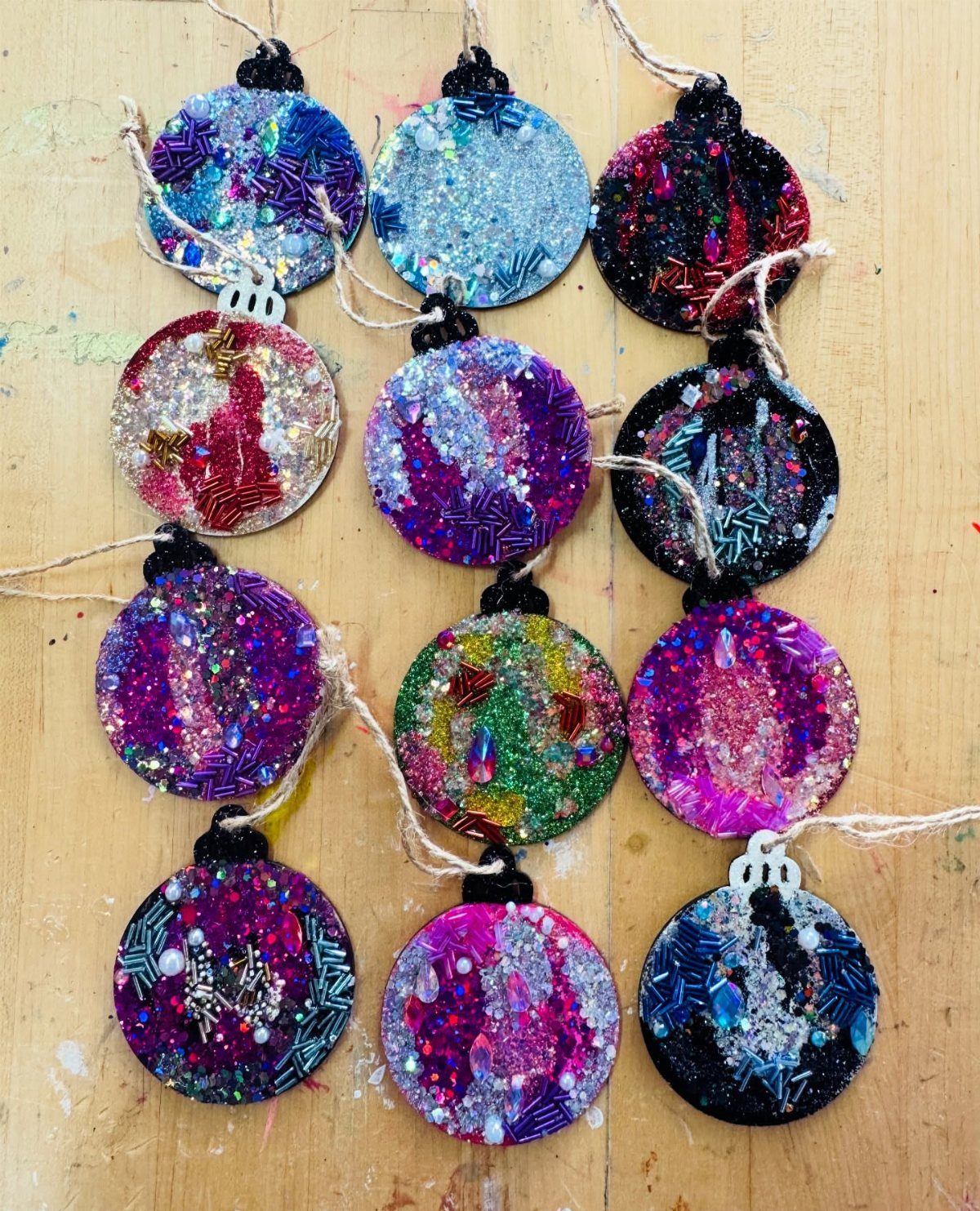 Glitter DIY Ornaments to Make 5 Ways 2 Twelve colorful glitter-covered flat ornaments arranged on a light wooden tabletop.