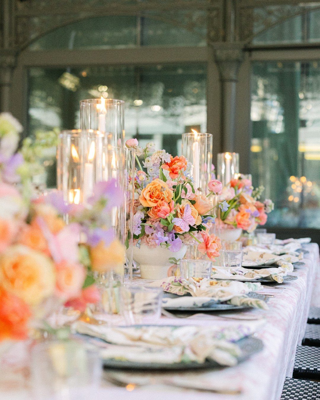 An elegantly decorated outdoor dining table featuring colorful floral arrangements and tall glass candle holders.