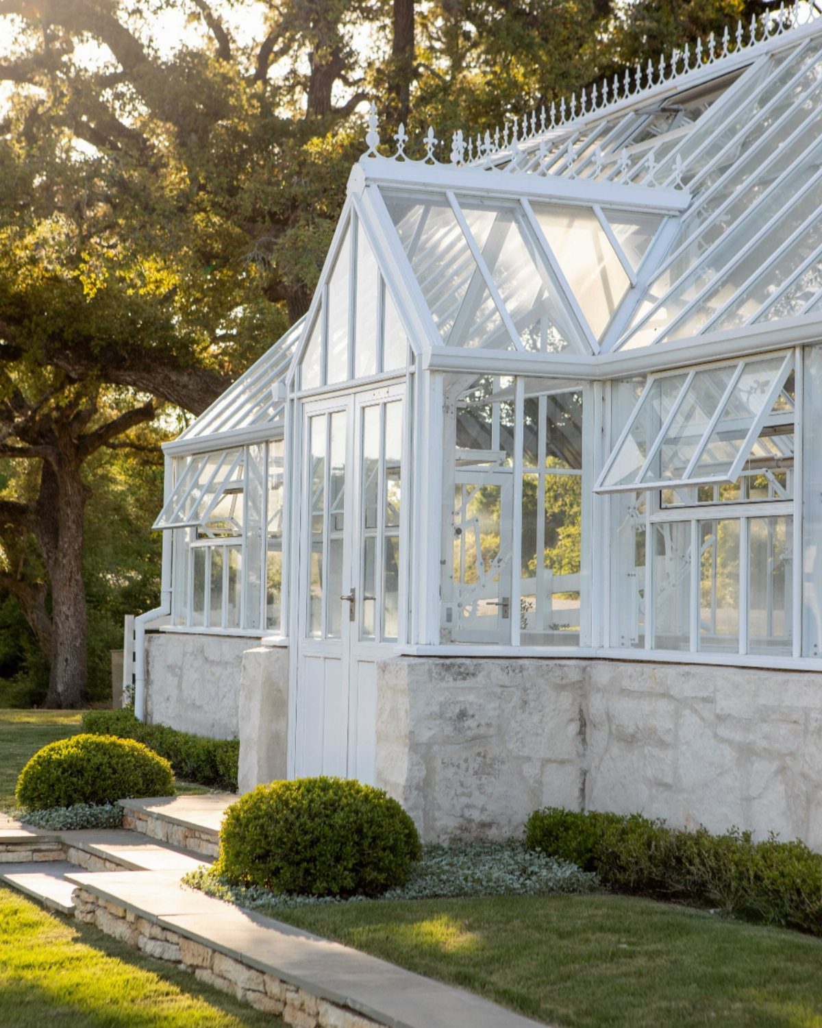 9 Greenhouse Garden Ideas for Every Size and Budget 4 A large white elegant greenhouse with glass walls and a stone foundation in a garden.
