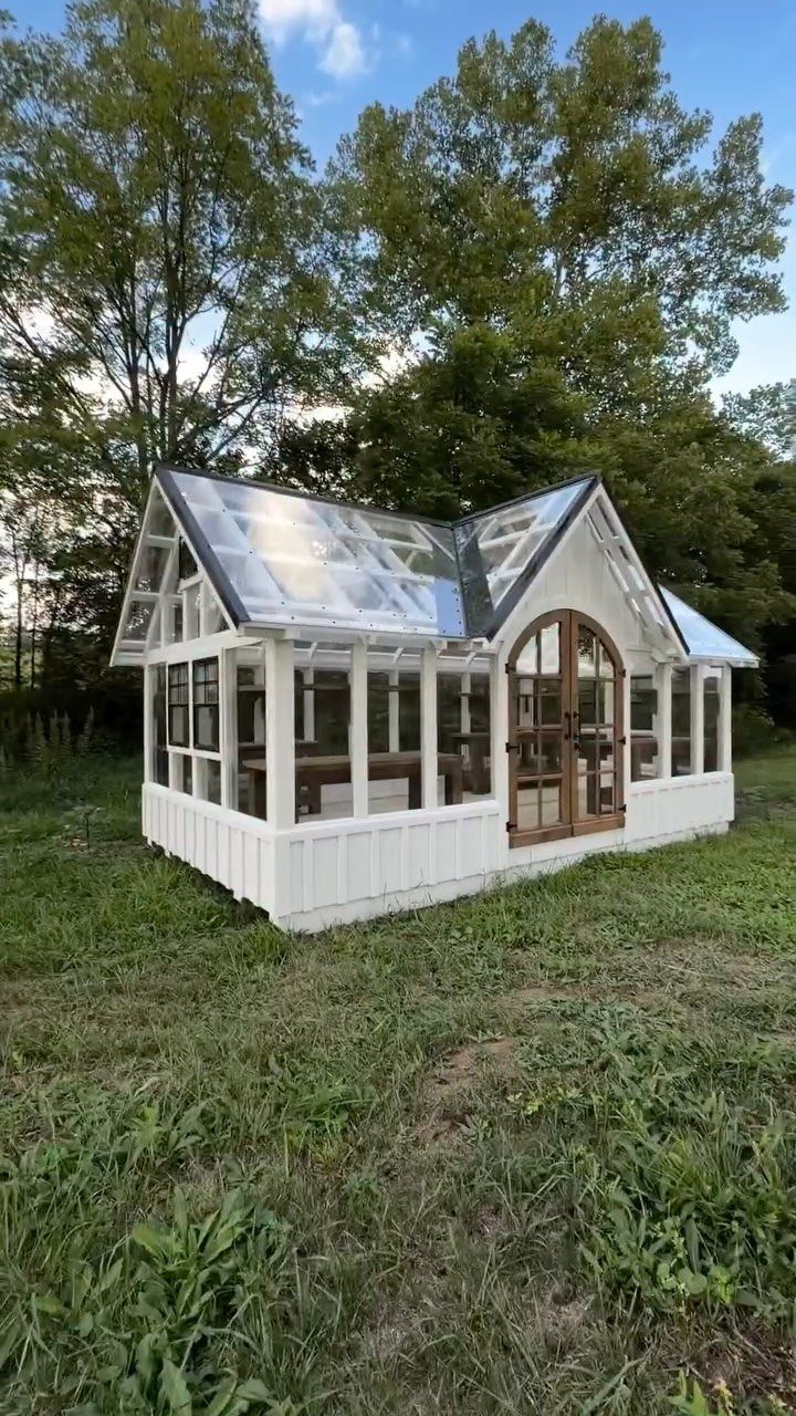 9 Greenhouse Garden Ideas for Every Size and Budget 2 A white wooden greenhouse containing a dining table and chairs in a grassy field.