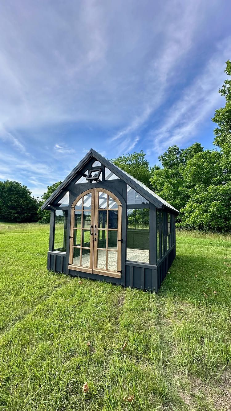 9 Greenhouse Garden Ideas for Every Size and Budget 5 A small black greenhouse with arched wooden doors sitting in a green grassy field.