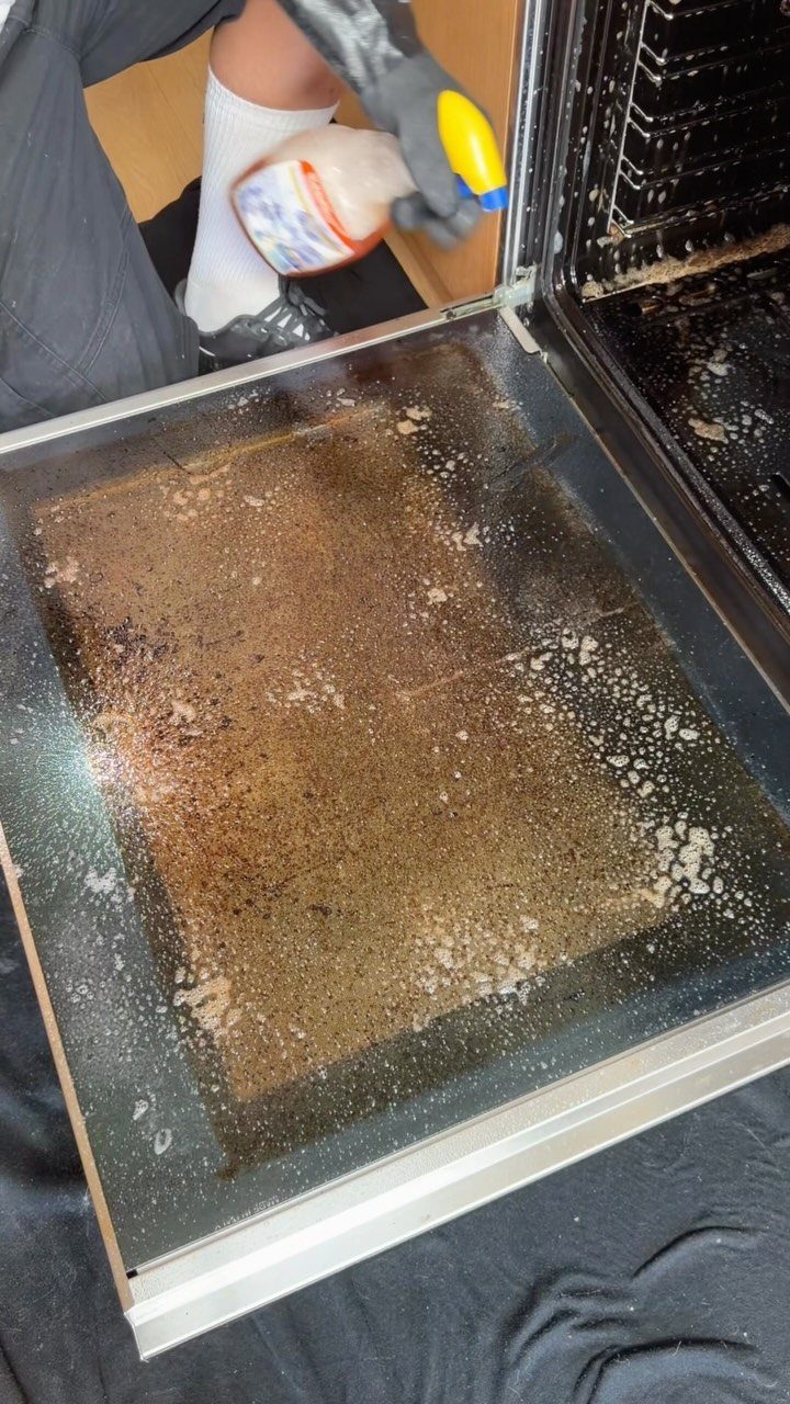 A person in black gloves sprays cleaner onto a dirty oven glass door.