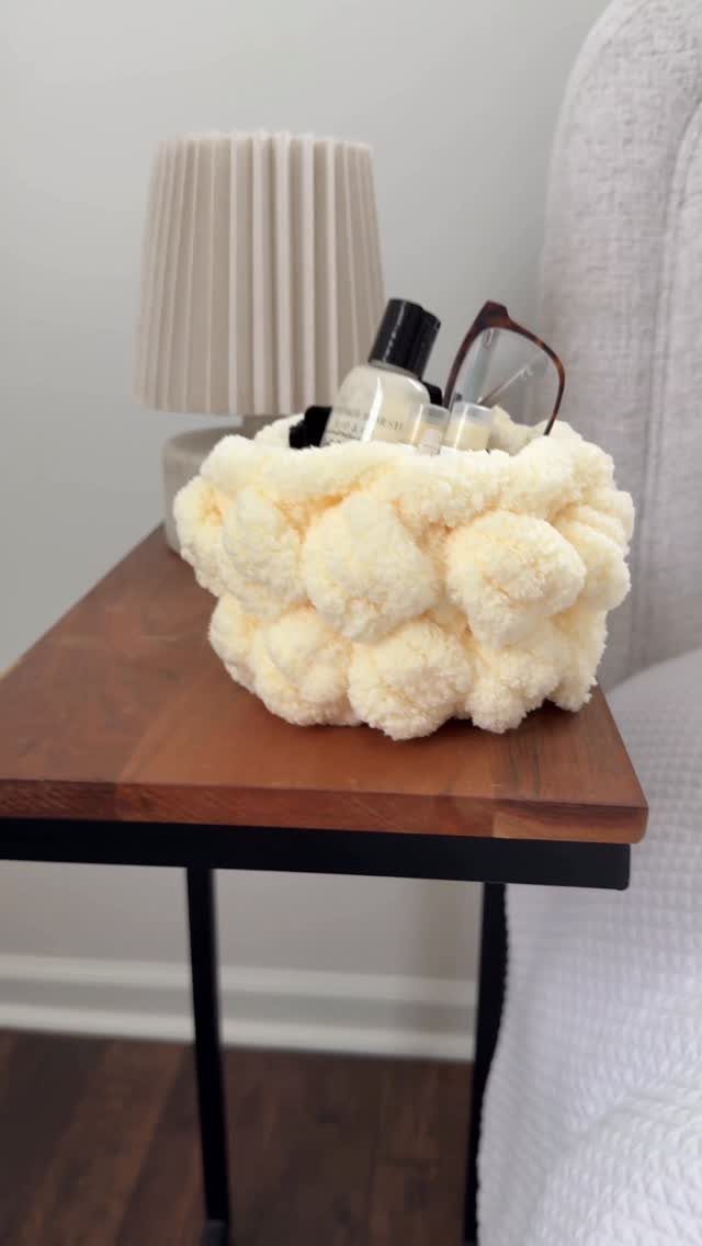 A textured cream pom-pom basket containing glasses and bottles on a wooden side table.