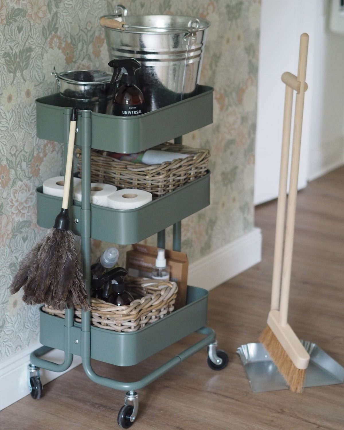 A green three-tiered rolling cart filled with cleaning supplies and buckets against a floral wallpapered wall.